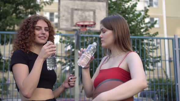 Two Happy Young Women Friends Drink Water After Training on the Street Basketball Court