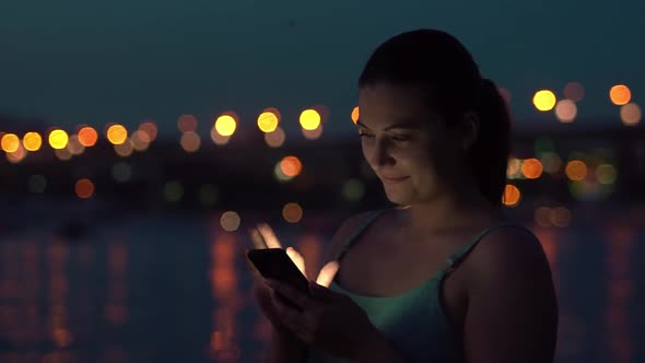 a Girl Uses a Smartphone While Walking Around the City at Night Having a Good Mood