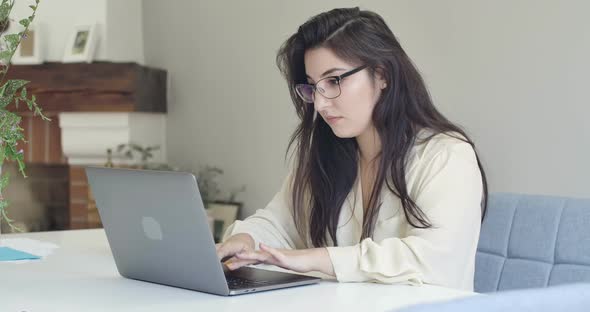 Portrait of Young Intelligent Caucasian Woman in Eyeglasses Typing on Laptop and Drinking Tea or