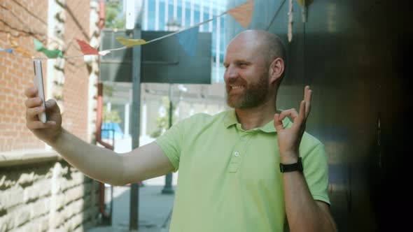 Stylish Young Man Taking Selfie with Smartphone Camera Showing Ok Hand Gesture Posing Outdoors