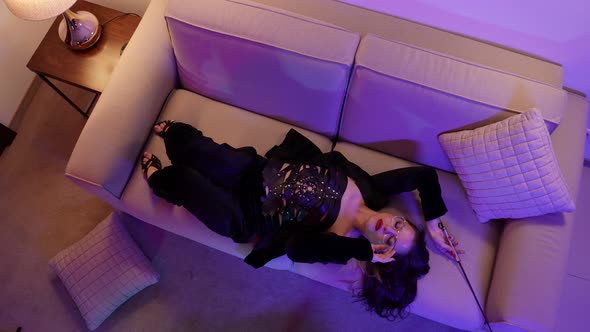 A Passionate Brunette Woman in Round Glasses Poses Lying on a Sofa in a Bright Room