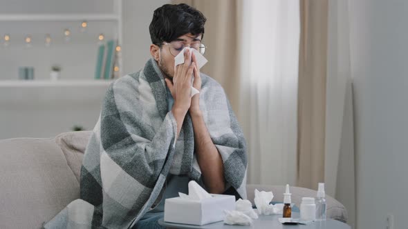 Young Sick Guy Arabic Man with Glasses Sits at Home Sofa Covered with Blanket Plaid Suffers From