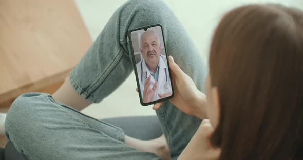 Female Medical Assistant Wears White Coat, Headset Video Calling Distant Patient on Smartphone