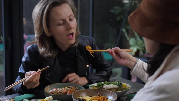 A Girl in a Hat Feeds Blonde Girl with Chopsticks. Sharing Your Food with the Loved Ones