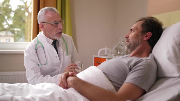 Aging Physician Examining Sick Male Lying in Ward