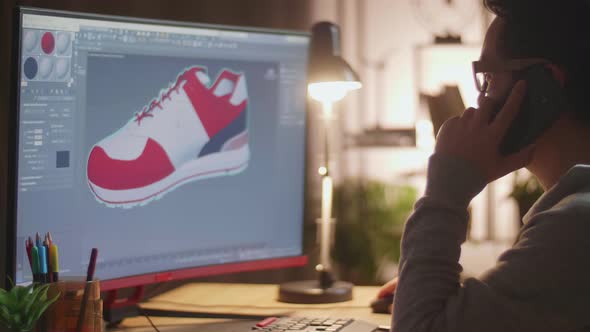 Asian Male Footwear Designer Talking On Smartphone While Designing Shoe On A Desktop At Home