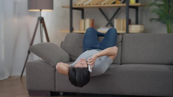 Young Asian Millennial Man Lying Upside Down on Couch Talking on Smartphone