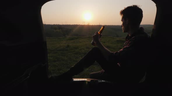 A Teenager Sits in the Trunk of a Car and Drinks Beer From a Bottle