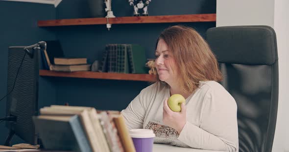 The Fat Woman is Working at Home at the Computer and is About to Eat an Apple