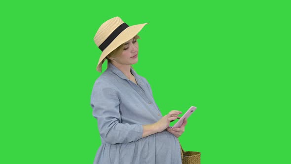 Happy Pregnant Woman in Blue Dress and Hat Using Her Smartphone on a Green Screen, Chroma Key.