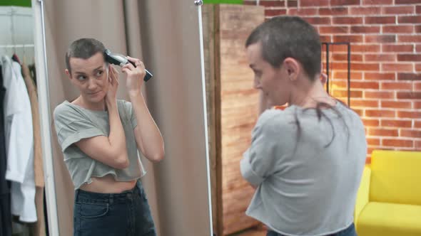 Shorthaired Woman Shaves Hair with a Machine for Haircut Standing in Front of a Mirror in the Room