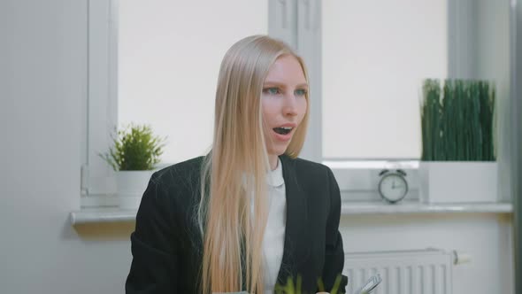 Woman Celebrating Success in Office. Elegant Blond Female Sitting at Workplace Holding in Hands