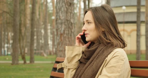 Young brunette woman talking on phone while sitting on a bench in autumn park wearing brown scarf.