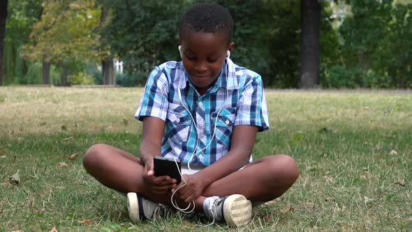 A Young  Black Boy Sits on Grass in a Park and Listens To Music on a Smartphone