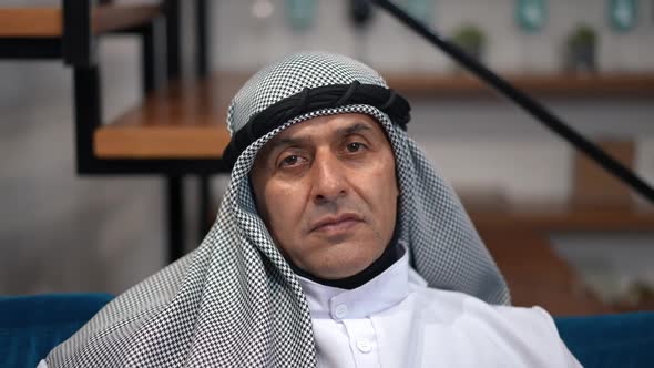 Zoom Out Confident Serious Middle Eastern Man Looking at Camera Sitting at Home in Living Room