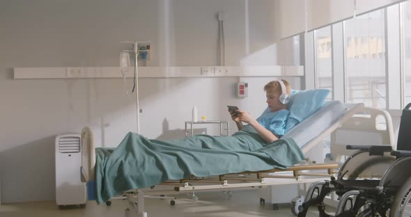 Sick Boy Relaxing in Hospital Bed with Digital Tablet and Headphones