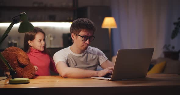 Tired Man Working on Laptop in Evening at Home and Cute Daughter Hugging Him