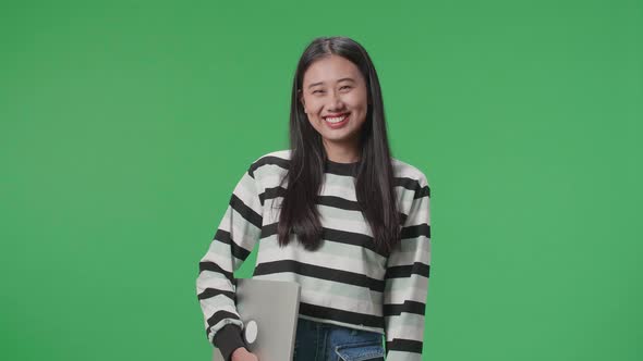 A Smiling Asian Woman Holding Computer While Standing In Front Of Green Screen Background