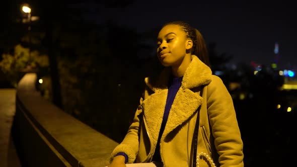 A Young Black Woman Waves at the Camera with a Smile As She Sits in a Park in an Urban Area at Night