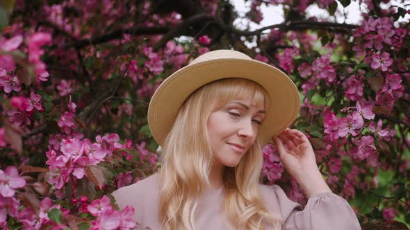 Portrait Romantic of a Blonde Woman in Purple Dress and Hat with Long Hair Standing By a Flowering