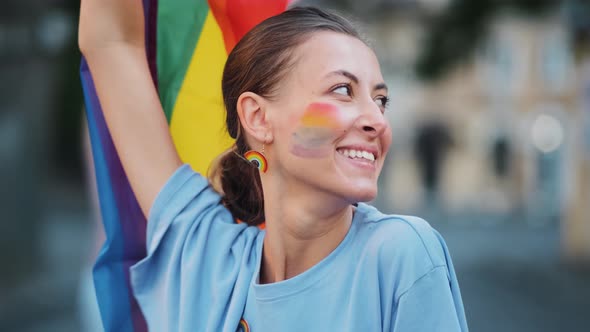 A smiling woman with colorful lgbt make-up waving colorful flag during pride gay parade