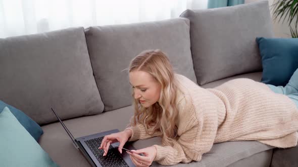 Blonde Girl Lying on the Couch Pays for Purchases on the Internet Using a Credit Card and Laptop