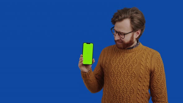 Handsome European Guy with Facial Hair and Glasses Looking and Pointing at His Smartphone with Green