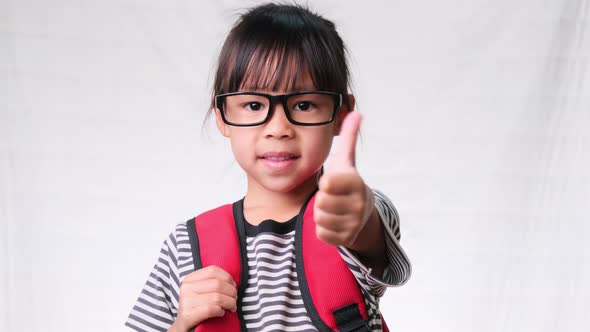 Happy schoolgirl wearing casual outfit with backpack showing thumbs up gesture on white