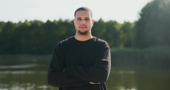 A Man in a Black Sweatshirt is Standing in Background of a Lake He Has His Arms Crossed Over Chest