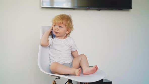 Little Son in Tshirt and Bloomers Sits on Chair with Phone