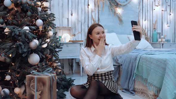 The Excited Young Woman Talks To Her Family with the Help of a Phone Sitting Next To the Christmas