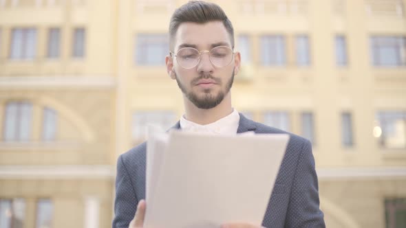 Portrait of Young Well-dressed Businessman in Glasses Standing in Front of High Building Reading