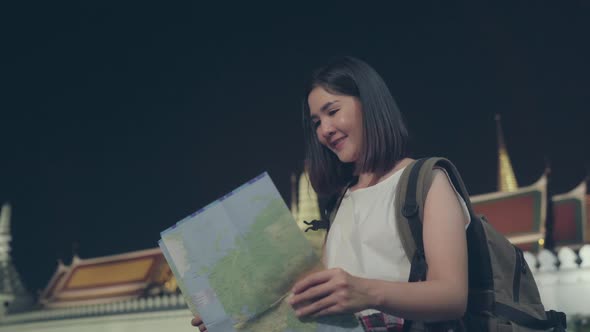 Young Asian tourist girl looking maps for direction places at night while walk along the street.