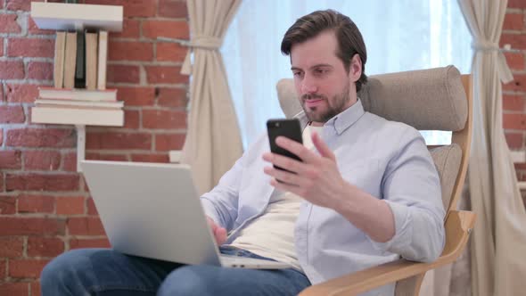Casual Young Man with Laptop Using Smartphone on Sofa