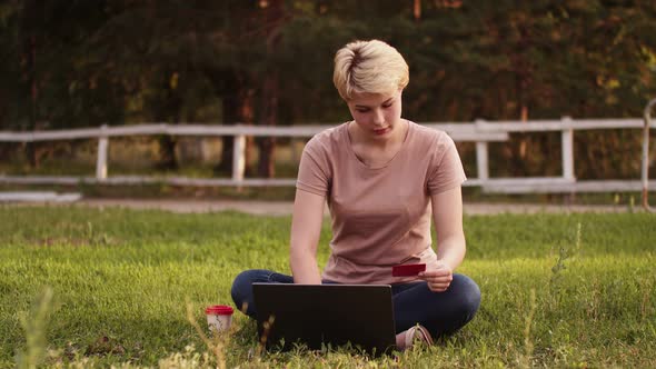 Confident woman is sitting on green lawn in park, holding credit card and using laptop.