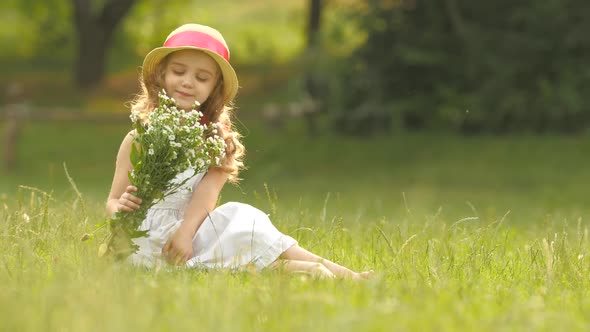 Baby Sits on a Glade in a Sarafan and Hat, Holds Wild Flowers and Smiles