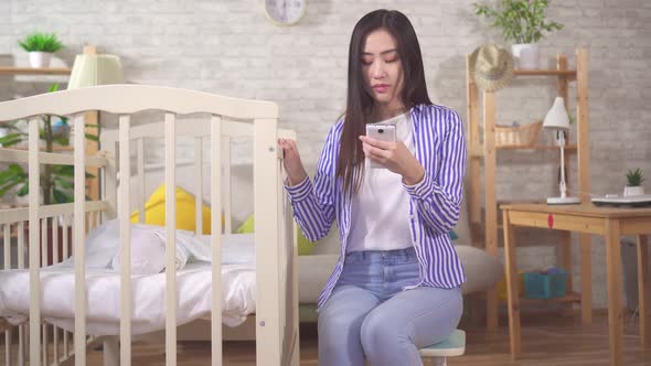 Young Asian Woman with a Smartphone Cradles a Baby in a Crib