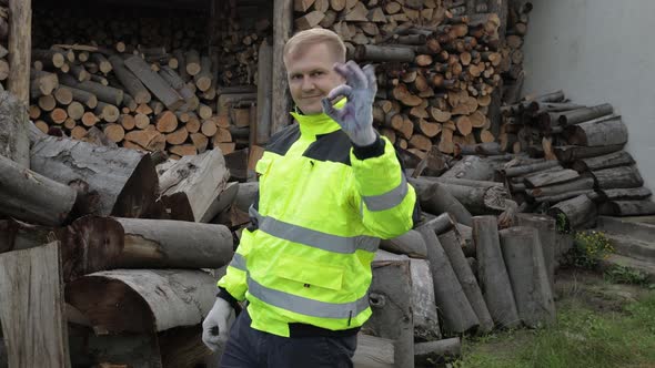 Lumberjack in Reflective Jacket. Man Woodcutter Show Ok Sign. Sawn Logs, Firewood Background