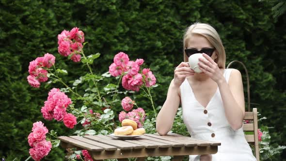 Blonde girl drink a coffee in garden with roses on background
