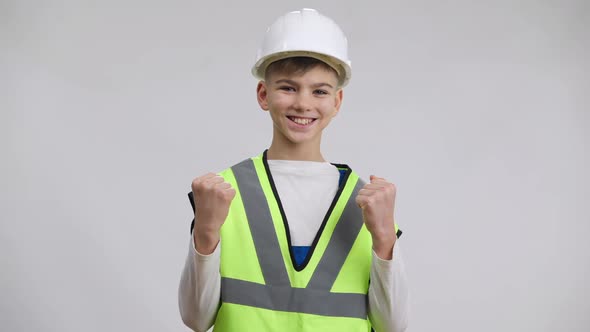 Positive Smiling Caucasian Boy in Hard Hat Making Victory Gesture Looking at Camera Standing at