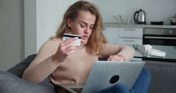 Blonde Woman is Making Online Payment Holding Bank Card Using Modern Laptop at Home Sitting on Sofa