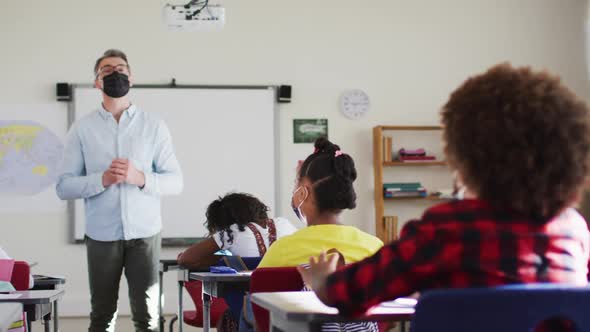 Diverse male teacher in classroom with children raising hands during lesson, all wearing face masks