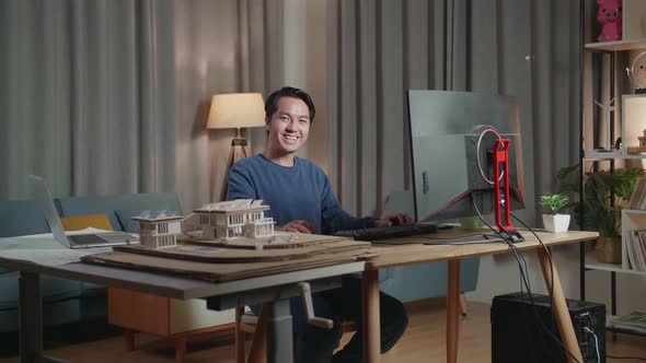 Asian Male Engineer With The House Model Smiling To The Camera While Working On A Desktop At Home
