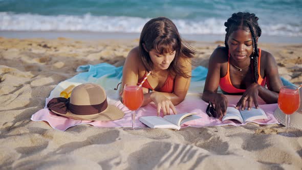 Two Girls Reading Books While Lying on Sandy Seashore