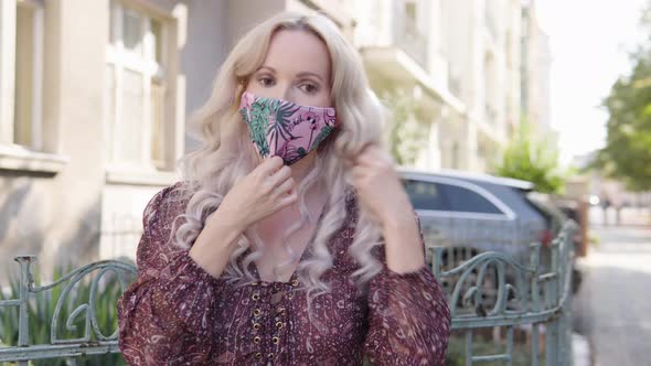 A Middleaged Caucasian Woman Puts on a Face Mask and Looks at the Camera in an Urban Area