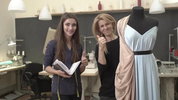 Blonde Caucasian Female Working on Mannequin with Azure Dress and Peachcolored Cloak with Woman in