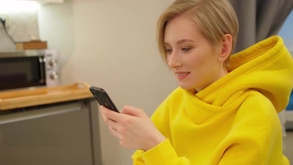 a Young Woman with a Short Haircut Sits in the Kitchen with a Smartphone in Her Hands and Smiles
