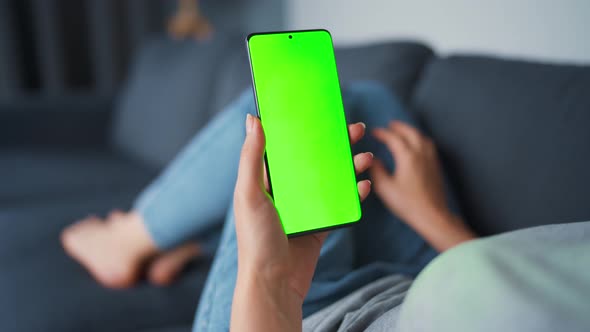 Woman at Home Lying on a Sofa and Using Smartphone with Green Mock-up Screen in Vertical Mode. Girl