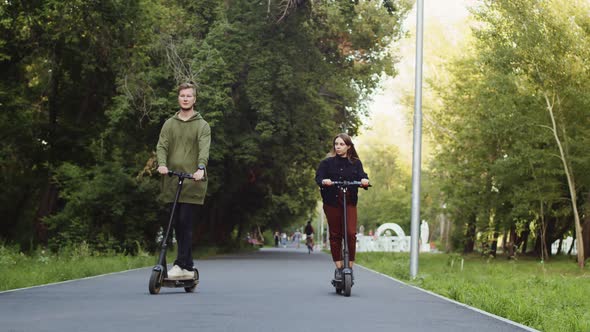 Man and Woman Ride Electric Scooters Along Alley in Public Park