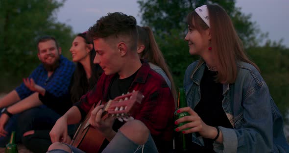 Hipster Man Playing on Acoustic Guitar and Singing Song with Friends Travelers at Big Bonfire at